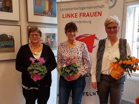 Ute Miething, Gabriele Brandt und Diana Bader stehen mit Blumensträußen in den Händen vor einem Roll-Up mit der Aufschrift "Landesarbeitsgemeinschaft LINKE Frauen Brandenburg"