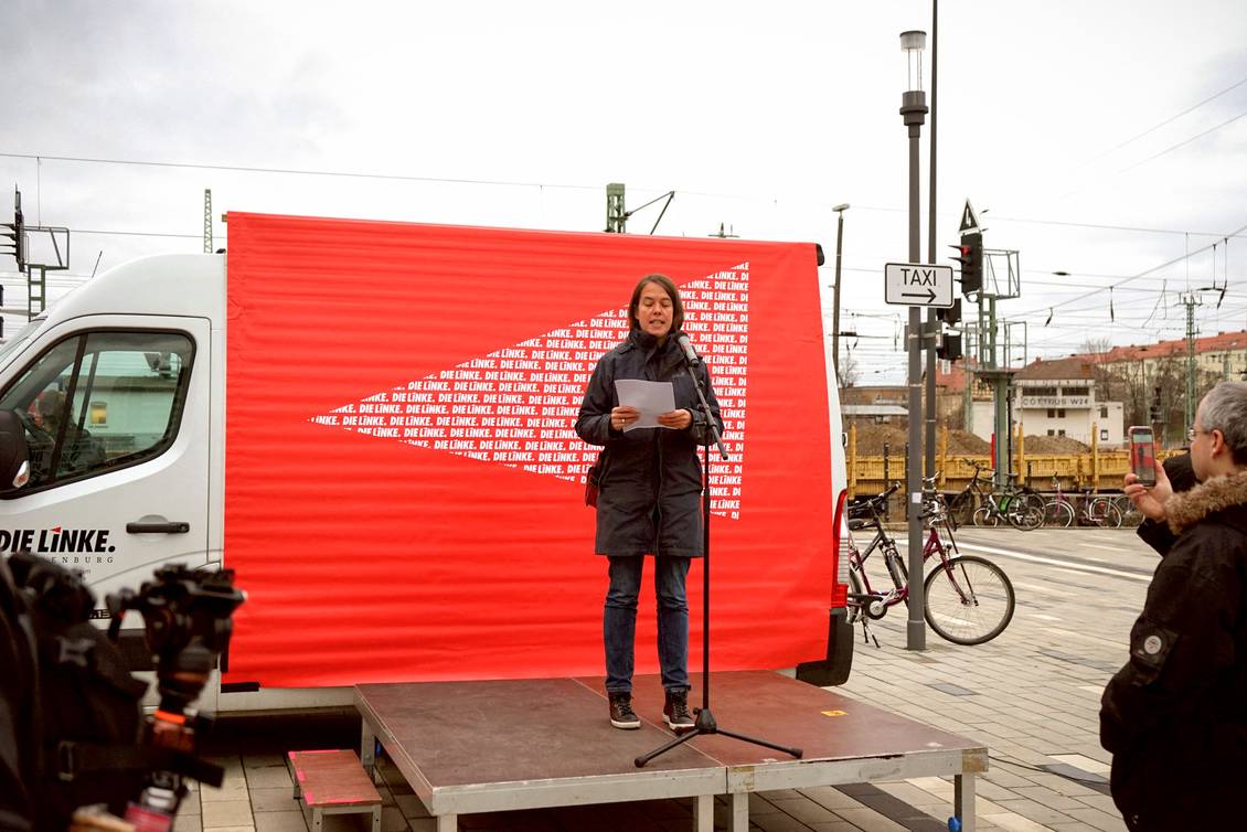 Christine Buchholz steht auf einer Bühne am Mikrofon vor einem Transporter, an dem ein Transparent mit LINKE-Logos hängt