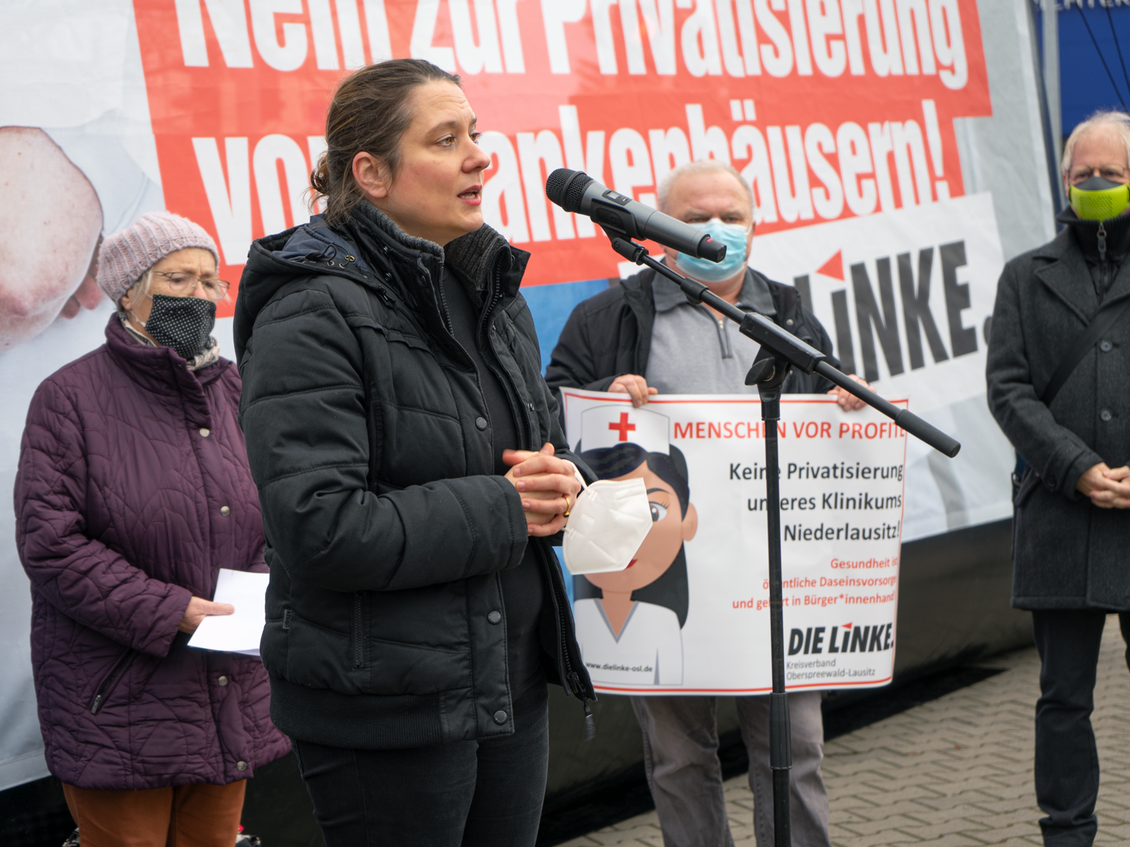 Anj Mayer spricht in ein Mikrofon, das an einem Mikrofonständer befestigt ist, neben ihr stehen Menschen mit Mund-Nase-Bedeckung