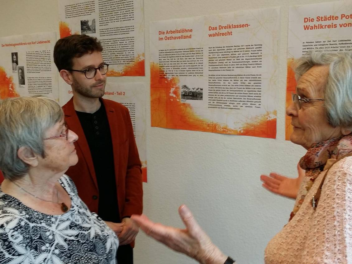 Tobias Bank und zwei ältere Damen stehen vor Ausstellungstafeln