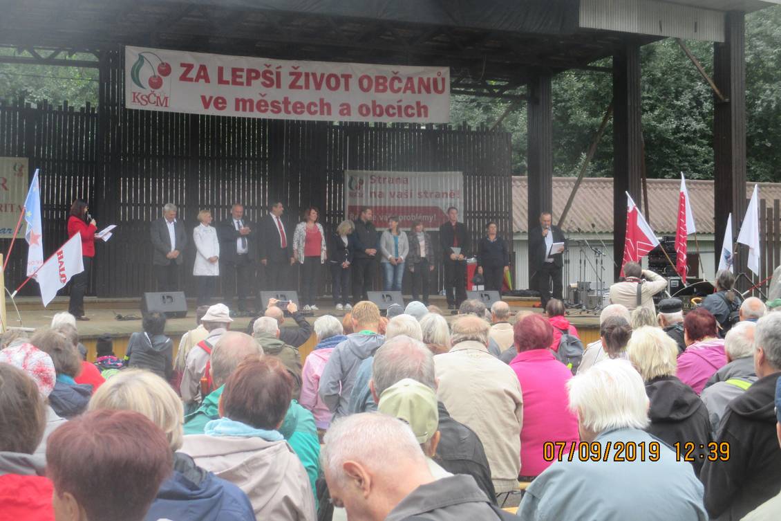 Eine Personengruppe steht auf einer Bühne, die mit Fahnen und einem Banner mit dem Logo der KSČM geschmückt ist