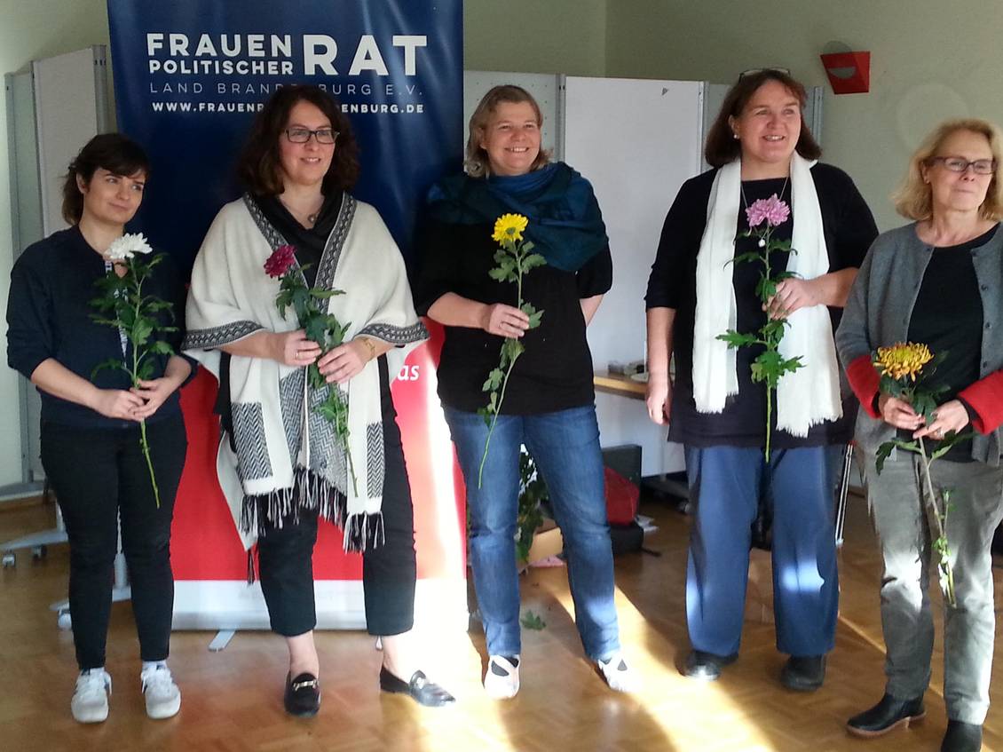Fünf Frauen mit je einer Blume in der Hand stehen vor einem Roll-Up mit der Aufschrift "Frauenpolitischer Rat"