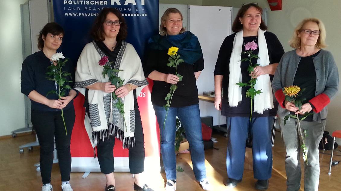 Fünf Frauen mit je einer Blume in der Hand stehen vor einem Roll-Up mit der Aufschrift "Frauenpolitischer Rat"