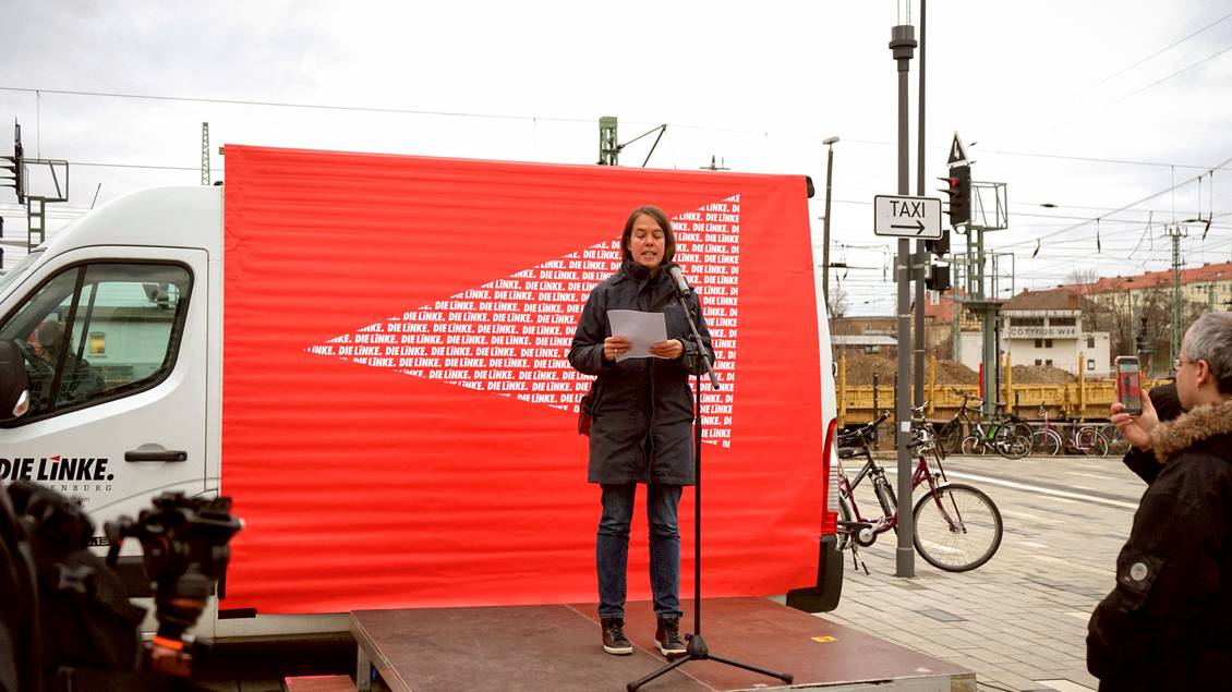 Christine Buchholz steht auf einer Bühne am Mikrofon vor einem Transporter, an dem ein Transparent mit LINKE-Logos hängt