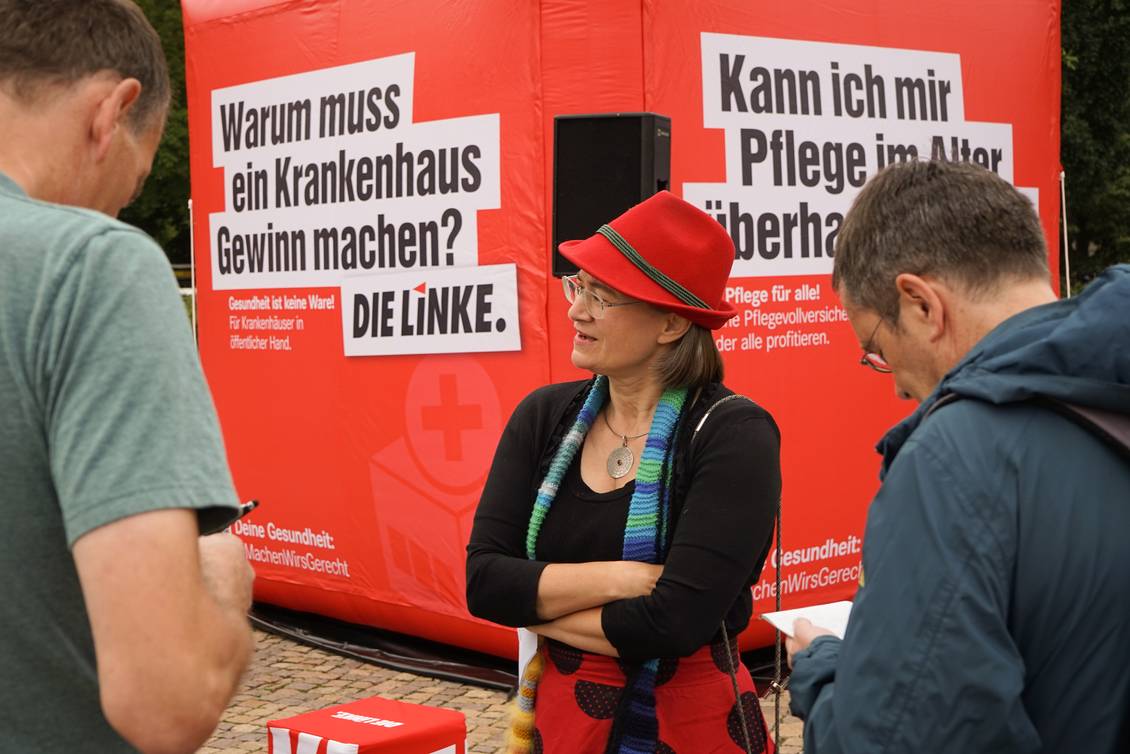 Anke Domscheit-Berg steht vor einem ein 3x3 Meter großen aufblasbaren roten Würfel mit dem Logo der LINKEN und verschiedenen Aussagen zu den Themen Pflege und Gesundheit und spricht mit zwei Personen