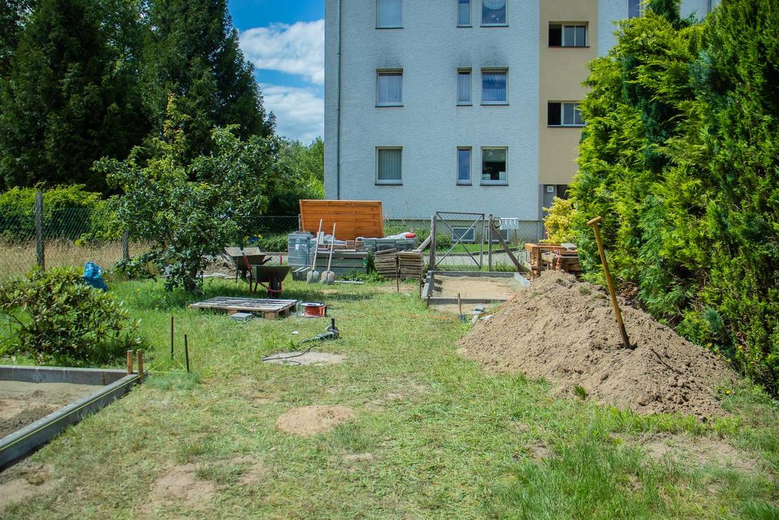 Gartengrundstück mit Bäumen und einem Sandhaufen, im Gras liegen Bauwerkzeuge