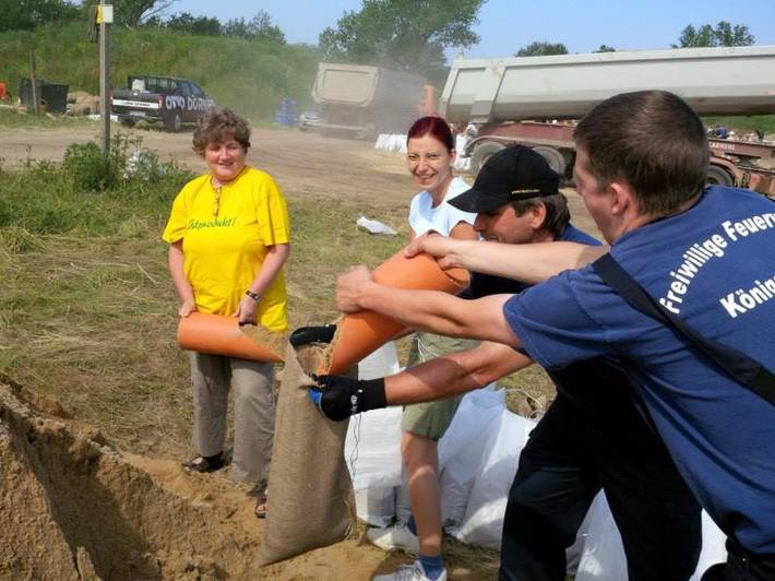Kirsten Tackmann, Diana Golze und zwei Herren mit T-Shirts der Freiweilligen Feuerwehr Königsberg füllen Sand in Sandsäcke