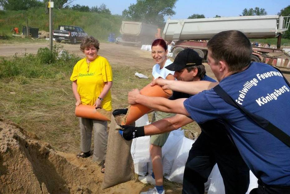 Kirsten Tackmann, Diana Golze und zwei Herren mit T-Shirts der Freiweilligen Feuerwehr Königsberg füllen Sand in Sandsäcke