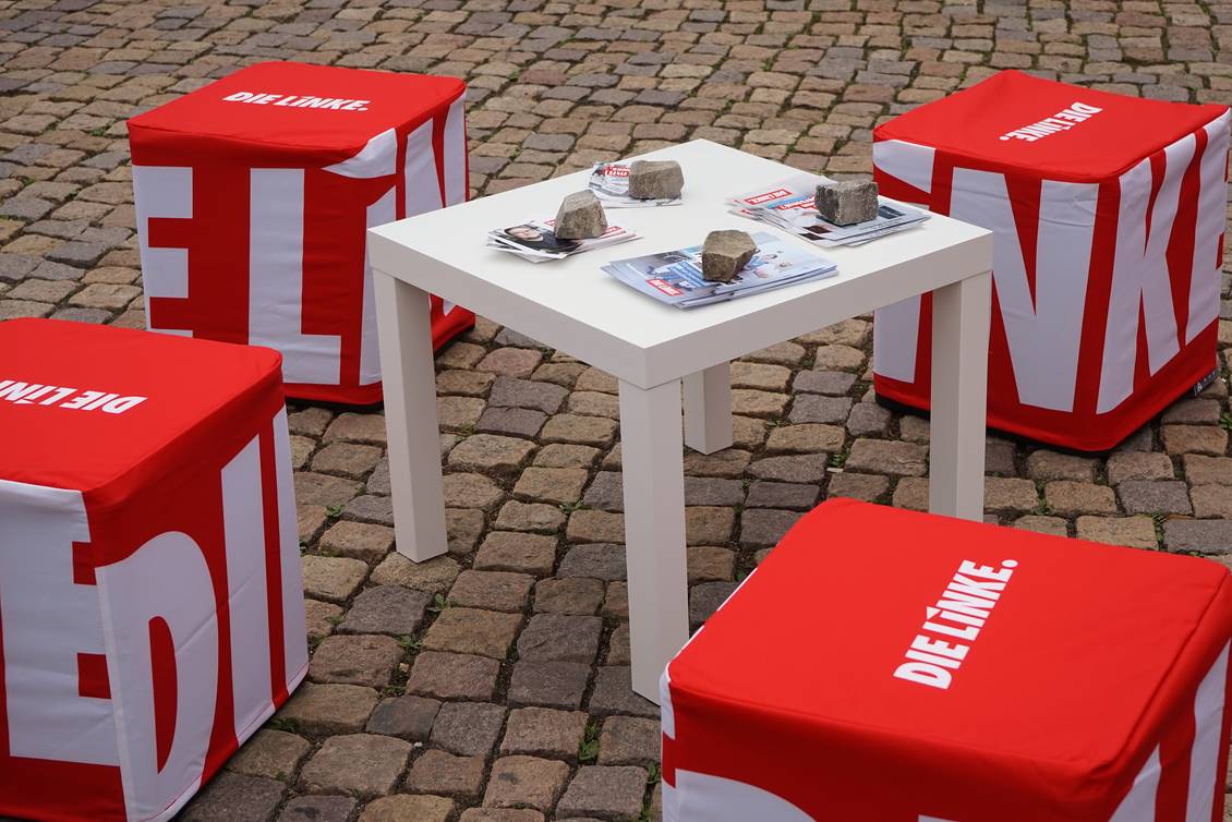 Ein weißer Beistelltisch, auf dem mehrere Stapel mit Flyern liegen, die mit Pflastersteinen beschwert wurden, um den Tisch herum stehen vier würfelförmige Hocker mit einer roten Husse mit dem Logo der LINKEN