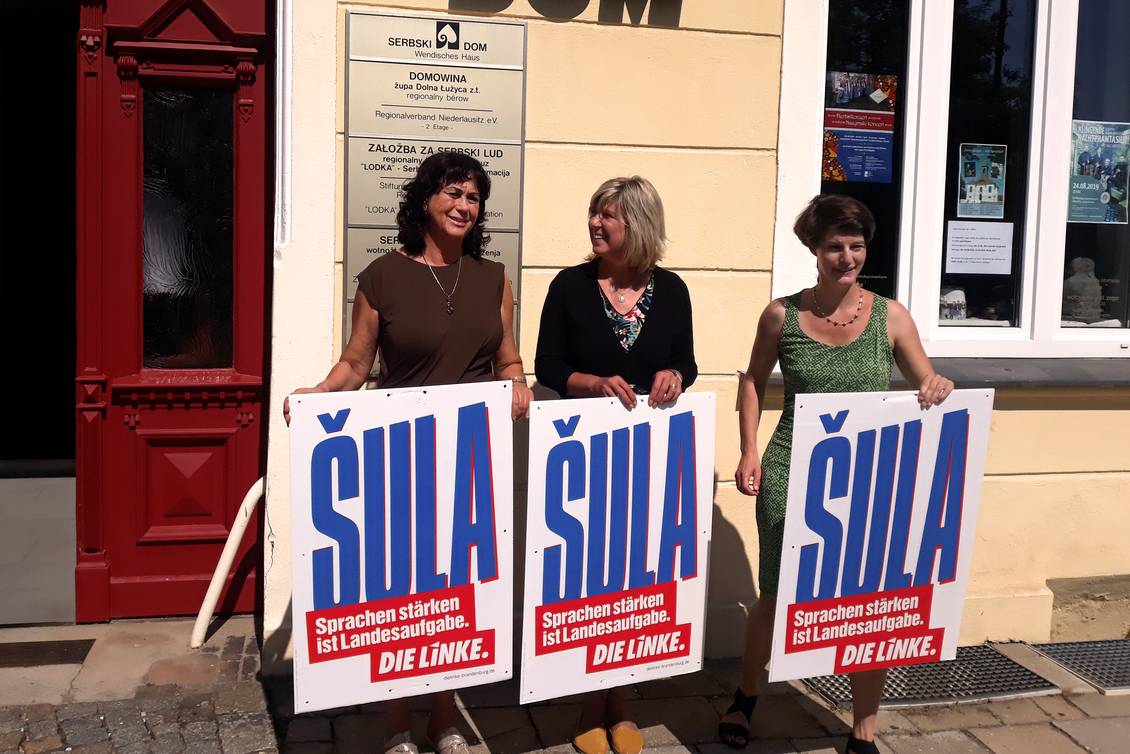 Birgit Kaufhold, Kathrin Dannenberg und Monika von der Lippe stehen vor dem Wendischen Haus in Cottbus und halten Wahlplakate hoch, auf denen "Šula" steht