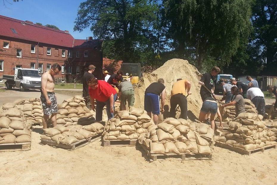 Mehrere Menschen füllen Sandsäcke mit Schaufeln, neben ihnen sind gefüllte Sandsäcke auf Europaletten gestapelt