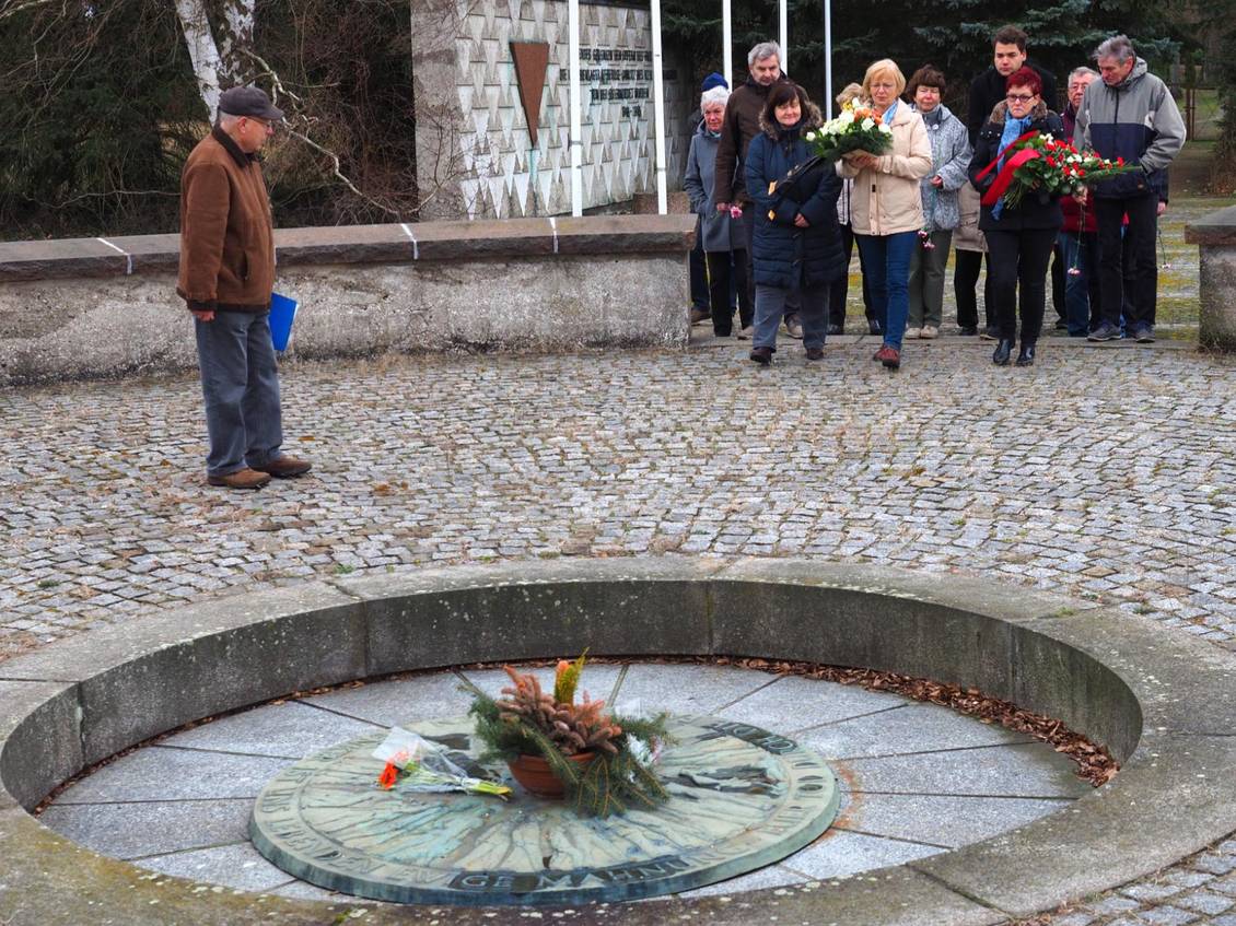 Eine Gruppe Menschen steht mit Gedenkkränzen vor dem Urnengrab in der Gedenkstätte für die Opfer des KZ Lieberose