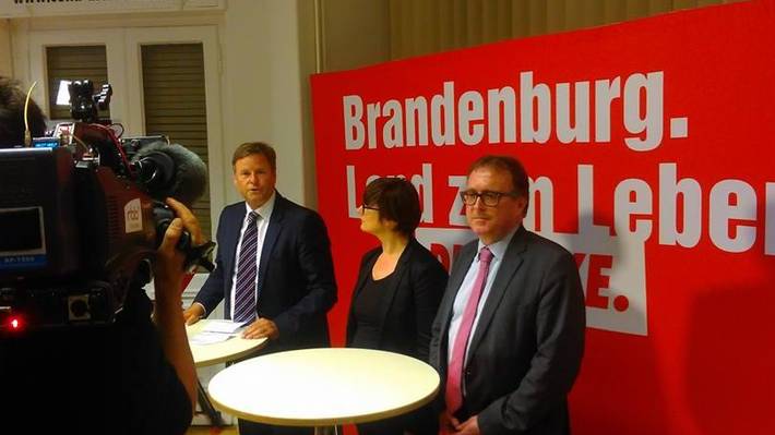 Christian Görke, Anja Mayer und Stefan Ludwig stehen an zwei Stehtischen vor einer roten Stellwand mit der Aufschrift "Brandenburg. Land zum Leben - DIE LINKE"