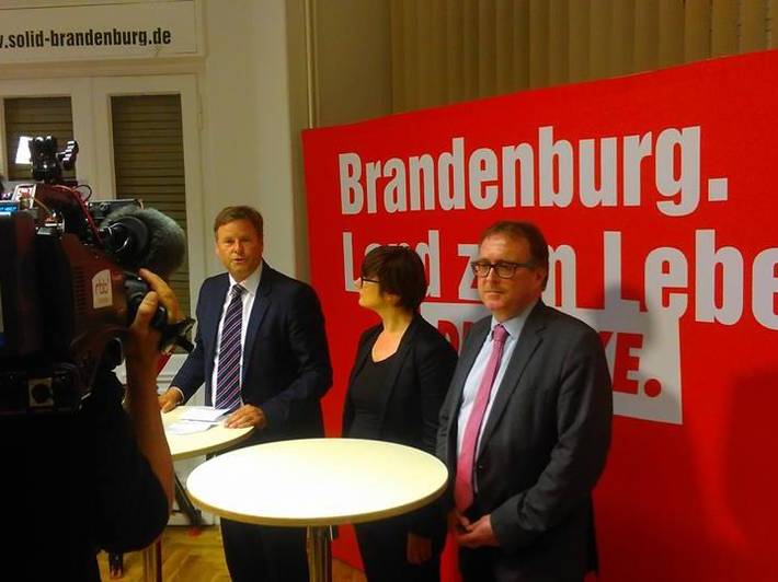 Christian Görke, Anja Mayer und Stefan Ludwig stehen an zwei Stehtischen vor einer roten Stellwand mit der Aufschrift "Brandenburg. Land zum Leben - DIE LINKE"
