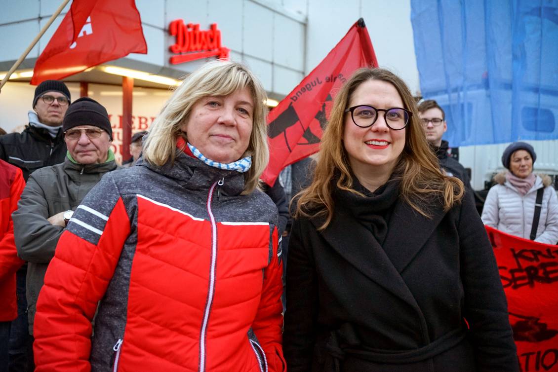 Kathrin Dannenberg und Anja Mayer stehen vor Demonstrierenden mit LINKE-Fahnen und Transparenten