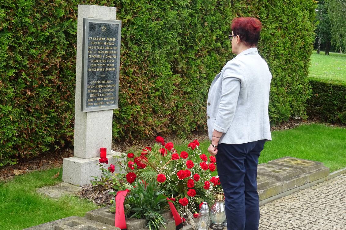 Monika Schömmel steht vor einer Stele mit einer Gedenktafel, vor der Blumen liegen