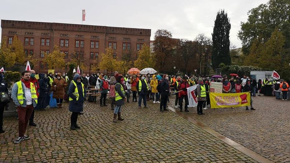 Menschen mit Warnwesten und Transparenten stehen auf einem Platz mit Kopfsteinpflaster