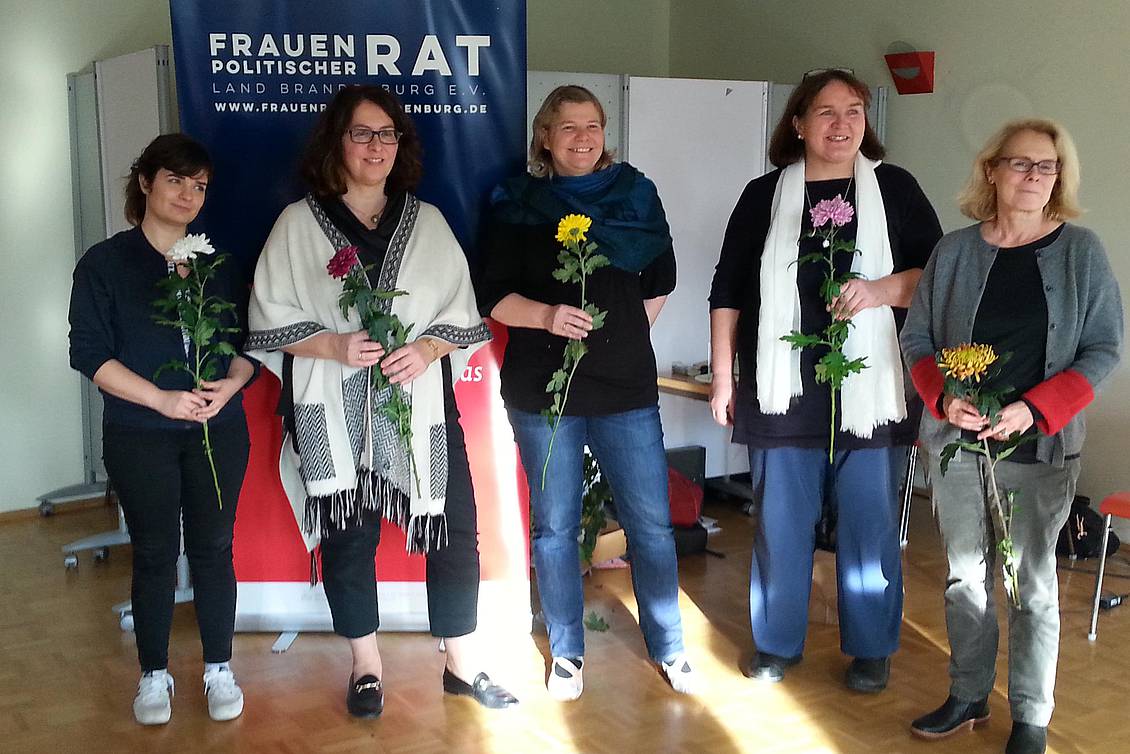 Fünf Frauen mit je einer Blume in der Hand stehen vor einem Roll-Up mit der Aufschrift "Frauenpolitischer Rat"