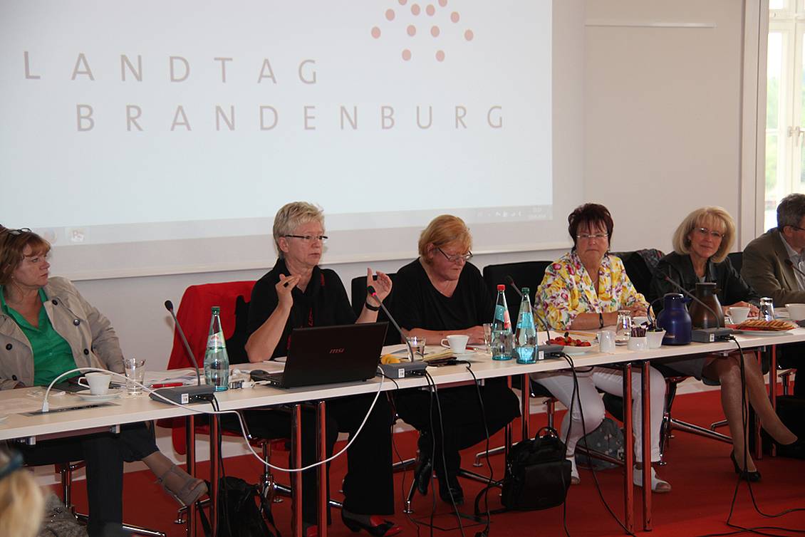 Konferenz in einem Besprechunsgraum mit Getränken auf den Tischen, an einem Tisch sitzt Anita Tack und spricht in ein Mikrofon, an der Wand eine Powerpoint-Folie auf der "Landtag Brandenburg" steht