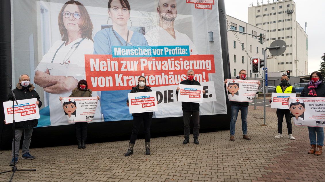 Mehrere Menschen stehen mit Abstand vor einer Großfläche mit dem Aufdruck "Menschen vor Profit: Nein zur Privatisierung von Krankenhäusern! DIE LINKE." und halten Schilder