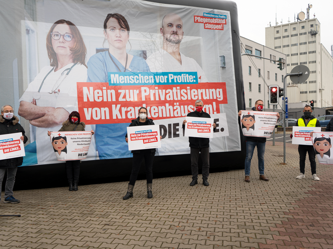Mehrere Menschen stehen mit Abstand vor einer Großfläche mit dem Aufdruck "Menschen vor Profit: Nein zur Privatisierung von Krankenhäusern! DIE LINKE." und halten Schilder
