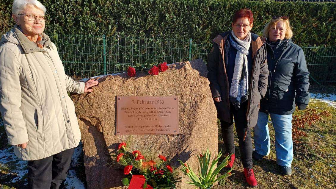 Mehrere Personen stehen am Gedenkstein, auf dem Blumen liegen