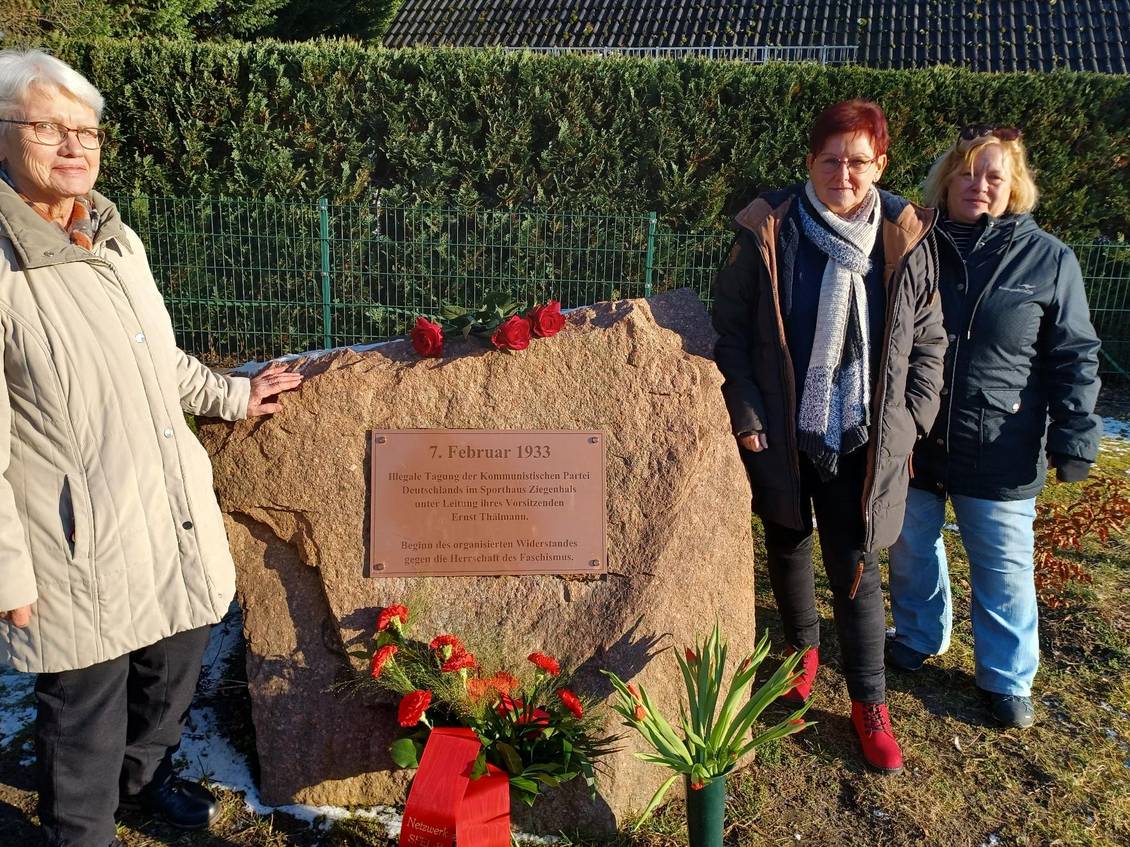 Mehrere Personen stehen am Gedenkstein, auf dem Blumen liegen