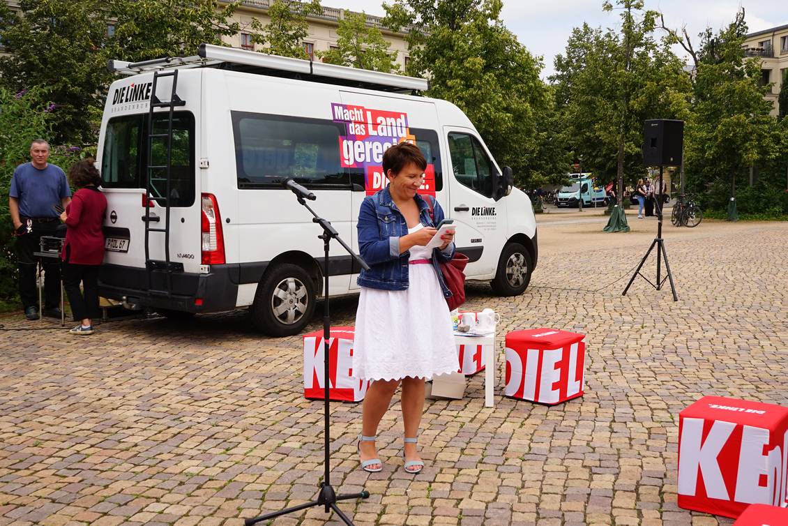 Katharina Slanina steht neben einem Mikrofonständer vor einem Kleinbus mit dem Logo der LINKEN