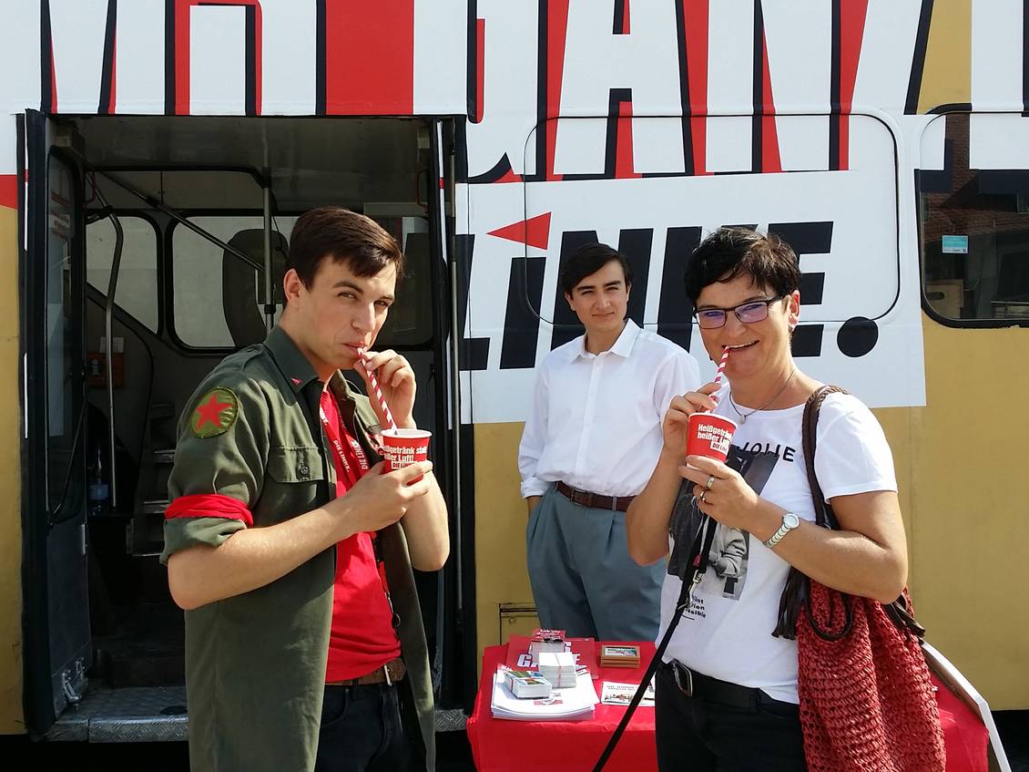 Aaron Birnbaum und Diana Bader stehen vor einem Doppeldecker-Bus und trinken mit Strohhalmen aus roten Pappbechern