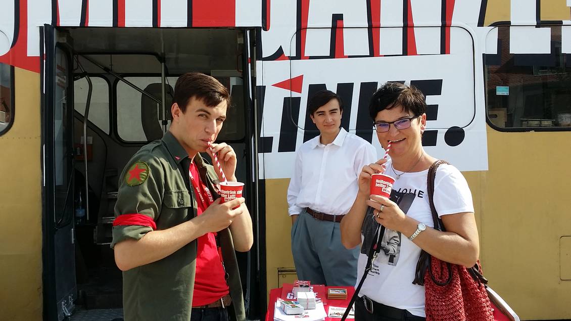 Aaron Birnbaum und Diana Bader stehen vor einem Doppeldecker-Bus und trinken mit Strohhalmen aus roten Pappbechern