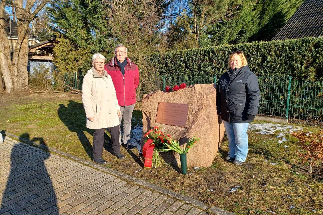 Mehrere Personen stehen am Gedenkstein, auf dem Blumen liegen