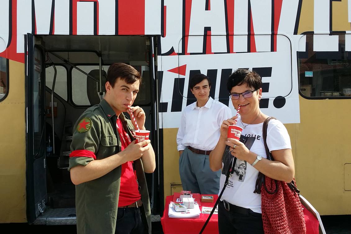 Aaron Birnbaum und Diana Bader stehen vor einem Doppeldecker-Bus und trinken mit Strohhalmen aus roten Pappbechern