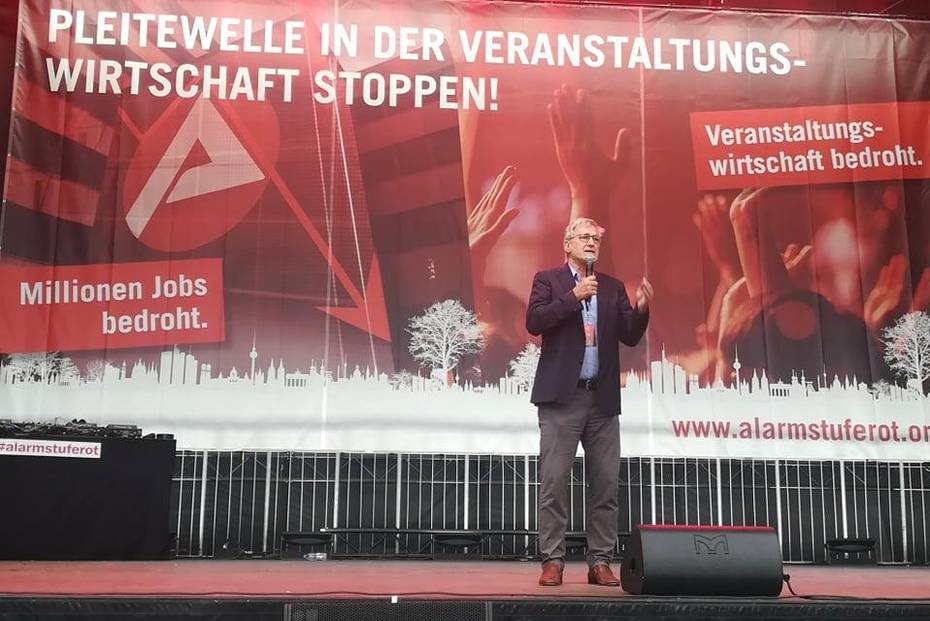 Bernd Riexinger steht auf einer Bühne mit einem Mikrofon in der Hand